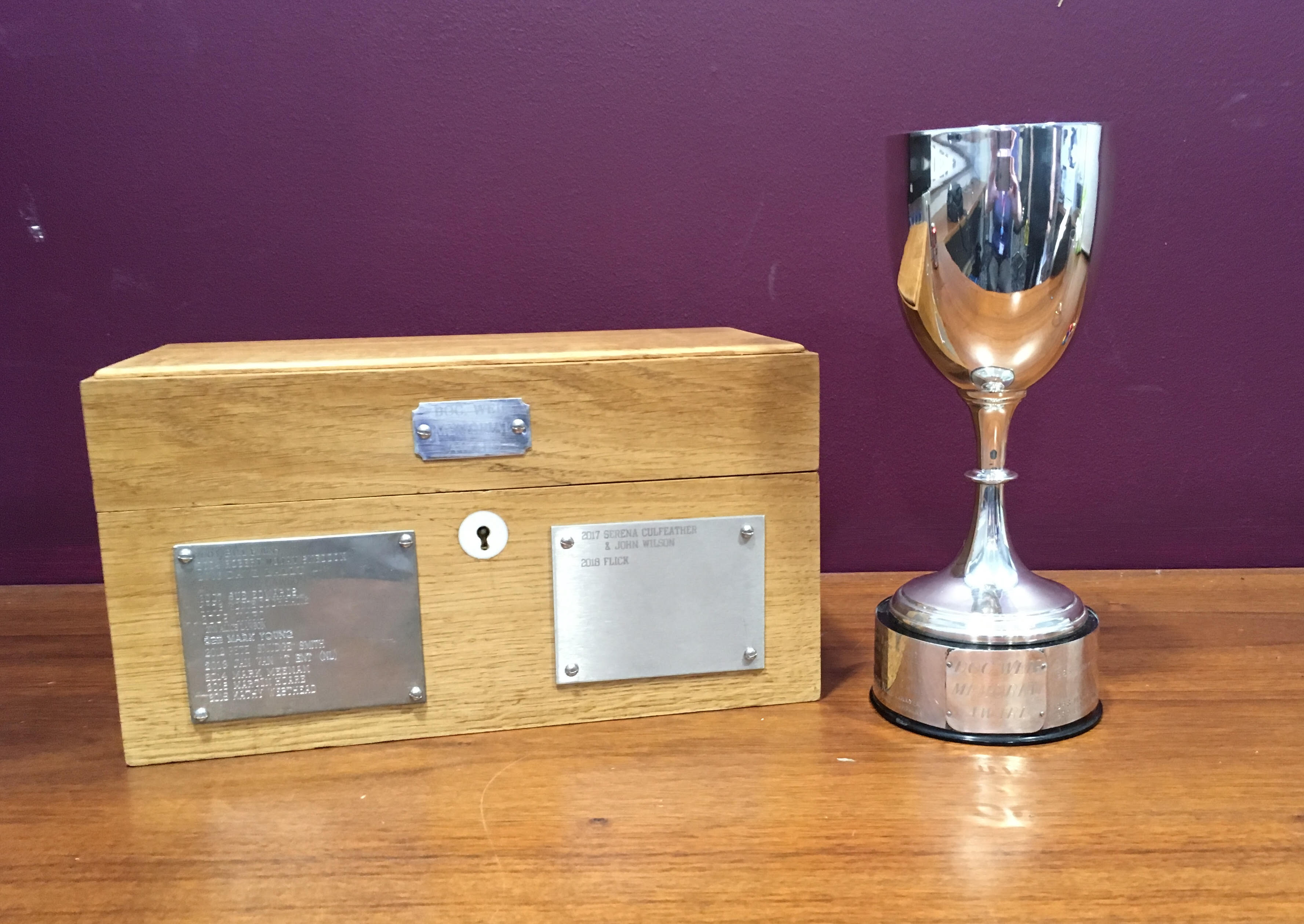 Photo of a wooden box next to a silver cup. On the front of the box are two plates with names of Doc Weir winners engraved on them.