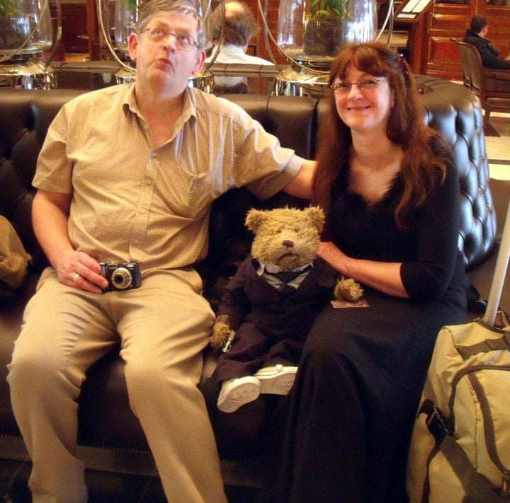 Photo of John Wilson, a white man, and Serena Culfeather, a white woman, seated on a hotel sofa with a besuited toy bear sitting between them.