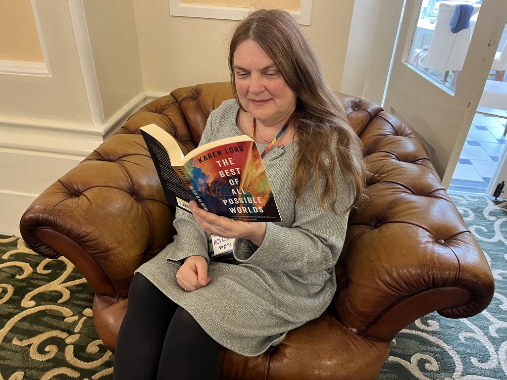 A photo of Virginia Preston reading Karen Lord's book "The Best of All Possible Worlds" while sitting on a brown leather armchair.