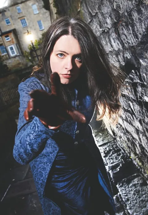 A photo of Emma Newman, a white woman, reaching out to the camera with a brown glove while dramatically lit.