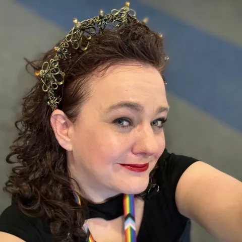 A woman smiling archly at the cameral.  She has long curly hair, is wearing makeup, an octopus tiara and a convention lanyard with the Pride flag