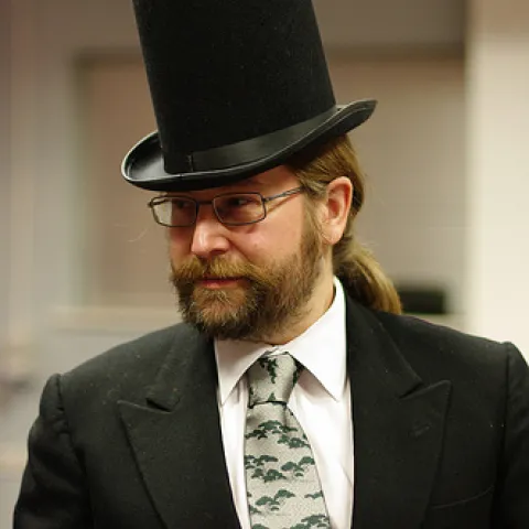 A bearded man in a dark suit and tie, wearing a large black top hat