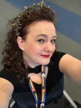 A woman smiling archly at the cameral.  She has long curly hair, is wearing makeup, an octopus tiara and a convention lanyard with the Pride flag