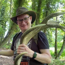 James stands in a forest holding an antler and wearing a hat