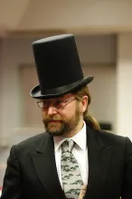 A bearded man in a dark suit and tie, wearing a large black top hat