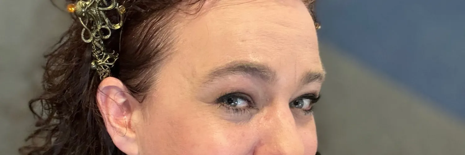 A woman smiling archly at the cameral.  She has long curly hair, is wearing makeup, an octopus tiara and a convention lanyard with the Pride flag