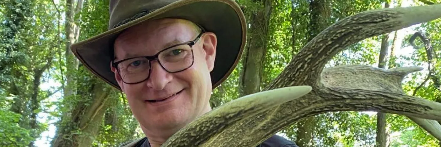 James stands in a forest holding an antler and wearing a hat