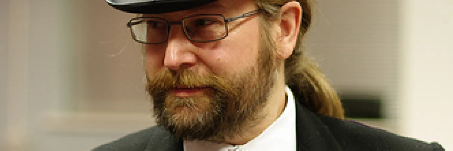 A bearded man in a dark suit and tie, wearing a large black top hat