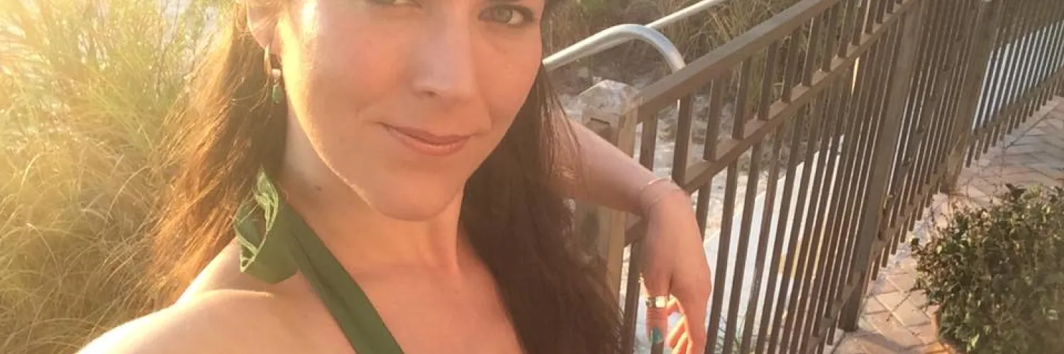 A woman with long dark hair, wearing a green halter-style dress, is smiling at the camera. She is outdoors in warm sunlight near a metal fence and greenery